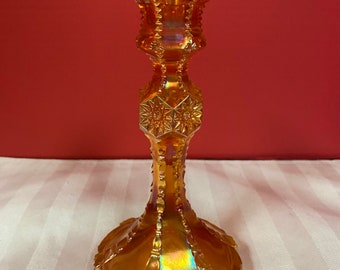 May include: An iridescent orange glass candlestick holder. The candlestick has a wide, ornate base, a slender stem with star-shaped details, and a flared top. The glass reflects a rainbow of colors.