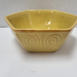 May include: A yellow ceramic bowl with a unique, hexagonal shape. The bowl features a decorative swirl pattern on the sides and a contrasting brown rim. This handcrafted bowl is perfect for serving food.