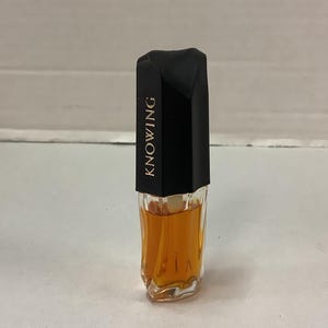 May include: A small, clear glass bottle of perfume with a black cap. The cap has the word "KNOWING" printed on it in gold letters.