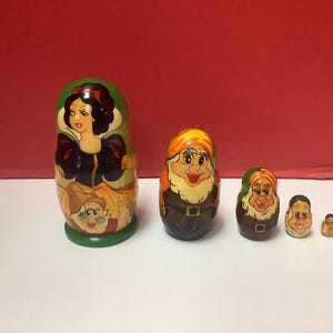 May include: A set of five nesting dolls featuring Snow White and the Seven Dwarfs. The largest doll depicts Snow White, while the smaller dolls represent the dwarfs. The dolls are painted in bright colors with detailed facial features.