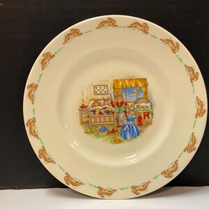 Puede incluir: Un plato de cerámica blanco con un borde verde y marrón que presenta un patrón repetitivo de conejos. El centro del plato tiene una ilustración colorida de conejos en un dormitorio.