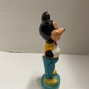 May include: A ceramic figurine of Mickey Mouse wearing a yellow shirt, blue jeans, and a red bow tie. He is standing on a blue and white base.