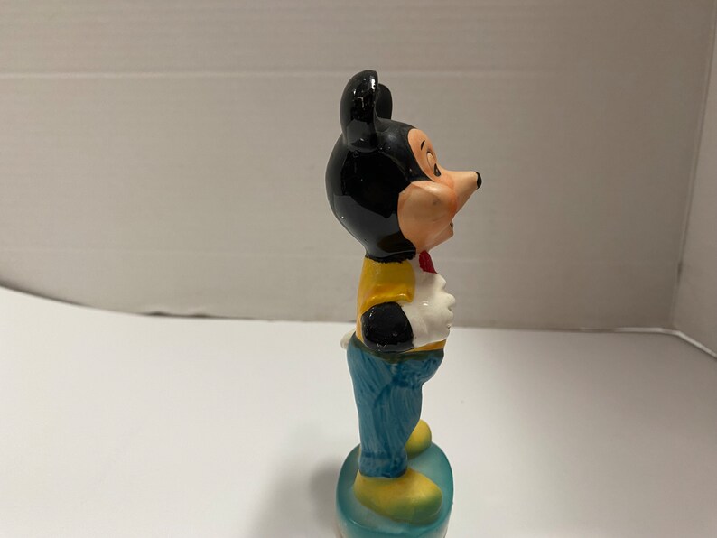 May include: A ceramic figurine of Mickey Mouse wearing a yellow shirt, blue jeans, and a red bow tie. He is standing on a blue and white base.