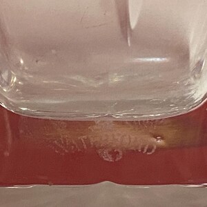 May include: Close-up of the bottom of a clear glass bottle with a red base. The bottle has a textured surface and the words "Cristal d'Arques" are etched into the bottom.