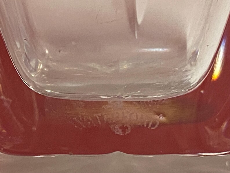 May include: Close-up of the bottom of a clear glass bottle with a red base. The bottle has a textured surface and the words "Cristal d'Arques" are etched into the bottom.