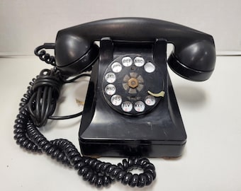 Könnte beinhalten: Ein schwarzes Wählscheiben-Telefon mit einem schwarzen Spiralkabel. Das Telefon hat eine schwarze Wählscheibe mit weißen Buchstaben und Zahlen. Das Telefon befindet sich auf einer weißen Oberfläche.