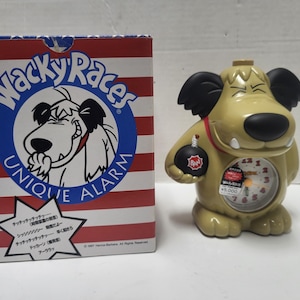 May include: A novelty alarm clock shaped like a cartoon dog, with a round clock face and a bomb in its paw. The clock is tan with black accents and a red collar. The box next to it has the "Wacky Races" logo.