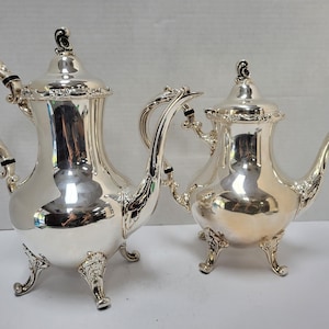 May include: Two polished silver-toned metal teapots featuring ornate, curved handles, decorative finials on the lids, and intricate scrollwork detailing on the spouts and feet. Both vessels have a reflective, smooth surface and a classic shape.