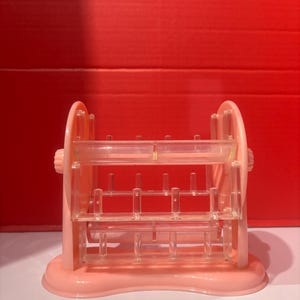 May include: A pink plastic thread spool holder with a rounded base and two side wheels. The holder has multiple horizontal bars with small pegs for holding spools of thread. The background is a solid red.