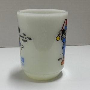 May include: White ceramic mug with a Mickey Mouse Club design featuring Mickey Mouse in a blue and red uniform holding a baton. The mug has the text "The Mickey Mouse Club" and the year "1955" printed on it.