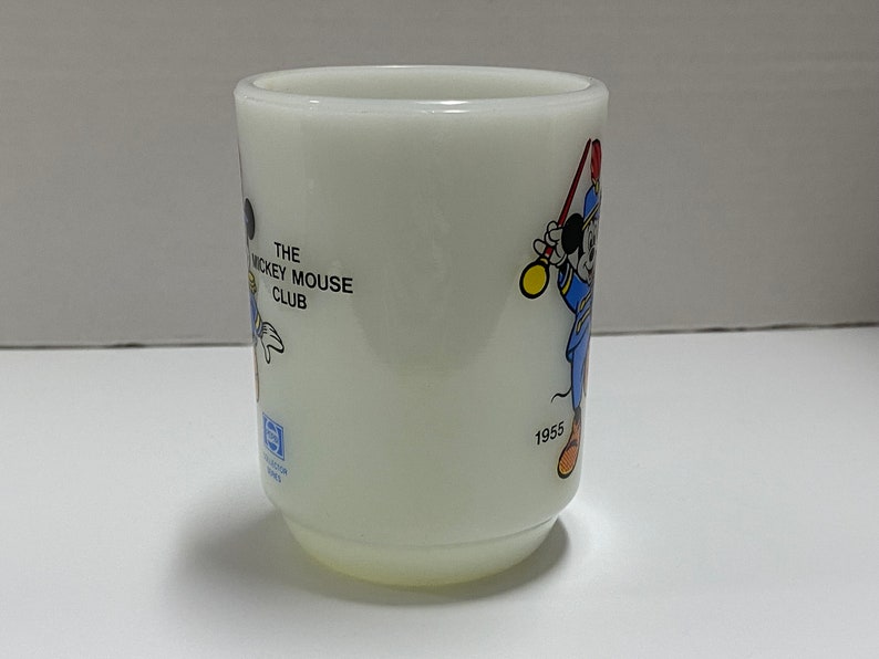 May include: White ceramic mug with a Mickey Mouse Club design featuring Mickey Mouse in a blue and red uniform holding a baton. The mug has the text "The Mickey Mouse Club" and the year "1955" printed on it.