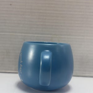 May include: A light blue ceramic mug with a rounded shape and a handle. The mug has the phrase "good day!" printed on the side in white script. The mug is sitting on a white surface.