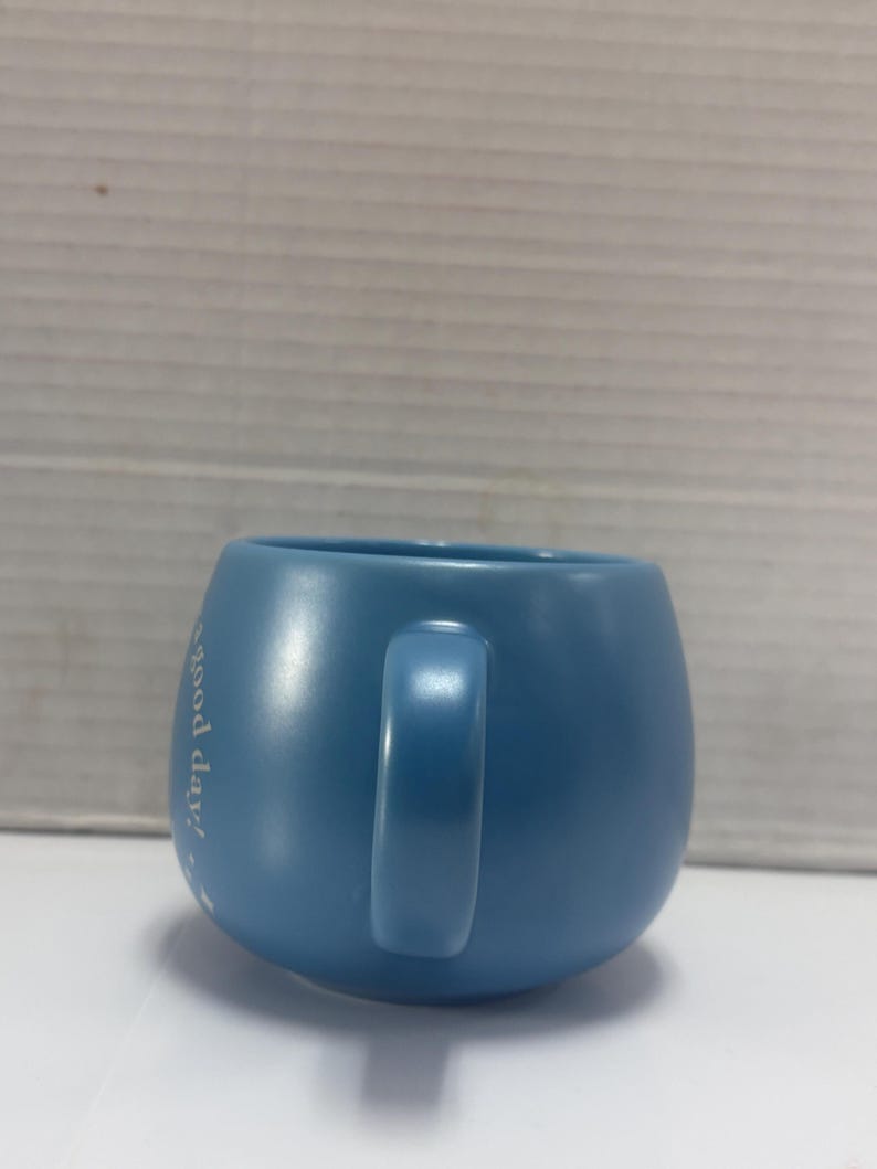 May include: A light blue ceramic mug with a rounded shape and a handle. The mug has the phrase "good day!" printed on the side in white script. The mug is sitting on a white surface.