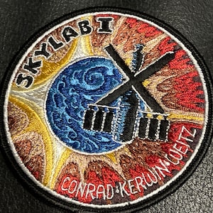 Puede incluir: Un parche bordado circular con una imagen en blanco y negro de la estación espacial Skylab, una imagen azul y blanca de la Tierra y el texto "SKYLAB I CONRAD-KERWIN-WEITZ".