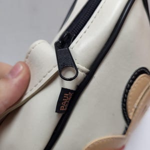 May include: Cream-colored rectangular bag with a black zipper and trim. The bag features a cartoon design with tan and red accents. The brand name "PAUL FRANK" is printed on a small label.