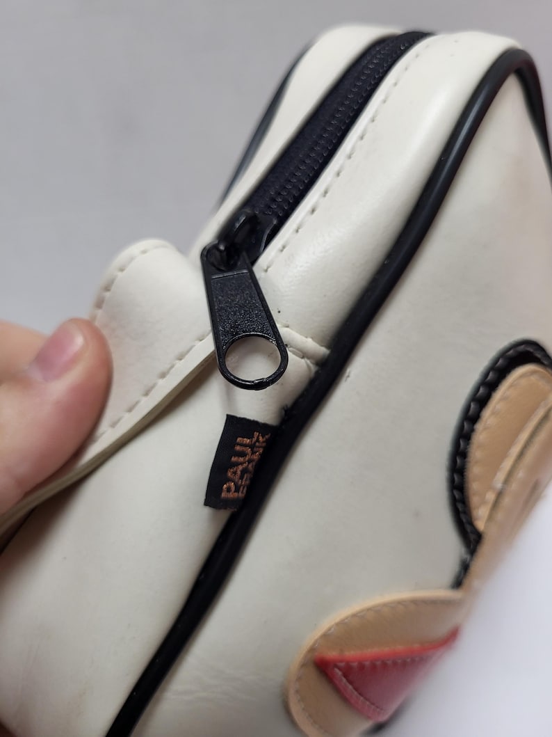 May include: Cream-colored rectangular bag with a black zipper and trim. The bag features a cartoon design with tan and red accents. The brand name "PAUL FRANK" is printed on a small label.