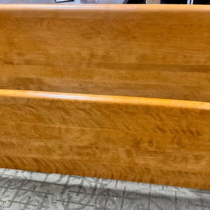 May include: A wooden bed frame set consisting of a tall headboard and a shorter footboard. Both pieces feature a smooth, light brown finish with a visible natural wood grain pattern and a gently curved top edge design.