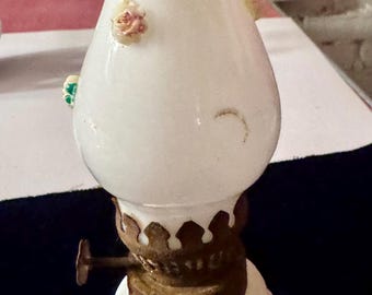 May include: A vintage white glass oil lamp with a ruffled top and floral accents. The lamp features a brass base and a decorative, textured design. Small, colorful flowers are affixed to the lamp's body.