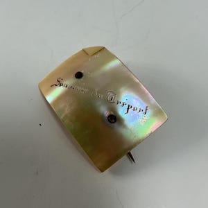 May include: Iridescent mother-of-pearl pin with the text "Souvenir du Treport" in script. The pin has a rectangular shape with a rounded top and a silver pin backing. Two small black dots are visible.