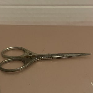 May include: A pair of silver-toned scissors with an ornate design on the handles. The scissors are closed and lying on a light brown surface.