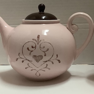 Op de afbeelding: Een roze keramische theeset met een theepot en twee mokken. De theepot heeft een bruin deksel en een decoratief ontwerp met een hart in het midden. De mokken en de theepot hebben een bijpassend ontwerp. De binnenkant van de mokken is bruin.
