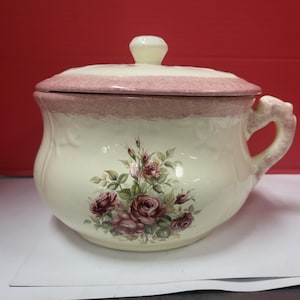 Pode incluir: Pote de cerâmica creme com tampa e alça. A tampa e a borda apresentam uma faixa rosa. O pote é decorado com um desenho floral de rosas em bordô e verde. A alça curva é detalhada.