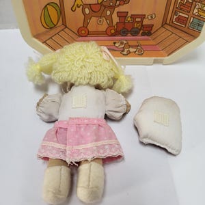 May include: A vintage doll with a yellow yarn wig, white top, and pink skirt with white lace trim. The doll has beige shoes and is next to a detached white piece. A toy house with illustrations of toys is in the background.