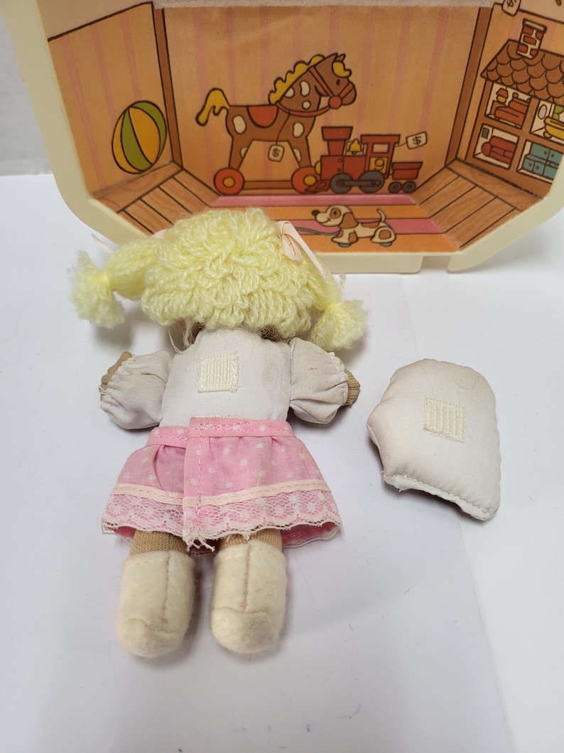 May include: A vintage doll with a yellow yarn wig, white top, and pink skirt with white lace trim. The doll has beige shoes and is next to a detached white piece. A toy house with illustrations of toys is in the background.