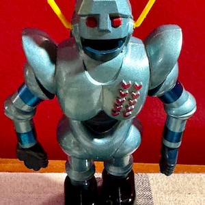 Puede incluir: Un juguete robot de plata con antenas amarillas, ojos rojos y botas negras. El robot tiene una cara sonriente y un patrón de rayas azules y blancas en los brazos.