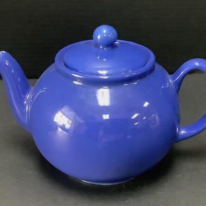 May include: A solid cobalt blue ceramic teapot with a rounded body, a curved spout, a matching lid with a small spherical knob, and a C-shaped handle. The teapot features a smooth, glossy finish that reflects light across its curved surface.