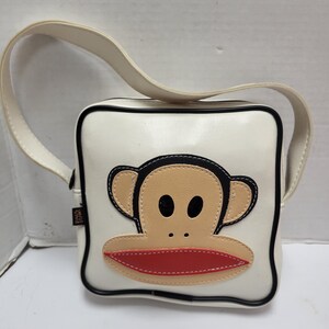 May include: White square crossbody bag with a black trim and a cream-colored strap. The bag features a large cartoon monkey face with tan, black, and red accents. The bag has a glossy finish.