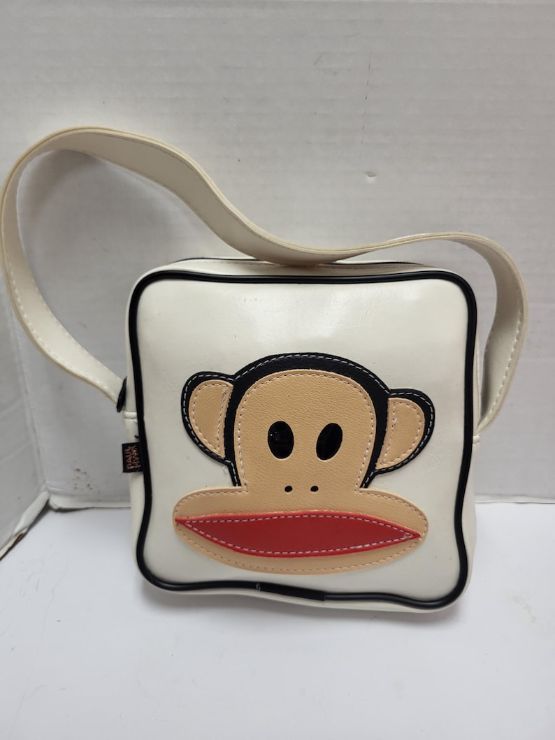 May include: White square crossbody bag with a black trim and a cream-colored strap. The bag features a large cartoon monkey face with tan, black, and red accents. The bag has a glossy finish.