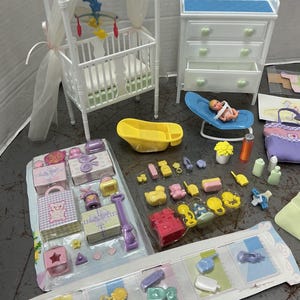 Może przedstawiać: Kolekcja miniaturowych mebli i akcesoriów dla niemowląt, w tym białe łóżeczko z baldachimem, komoda, wanienka dla dziecka i lalka w niebieskim krześle. Na zdjęciu znajdują się również pudełka na prezenty, zabawki i butelki.