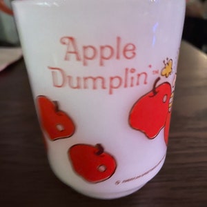 May include: White glass cup with the words "Apple Dumplin'" in red script. The cup features red apple illustrations with gold outlines. A vintage-style collectible cup.