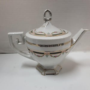 May include: White teapot with a floral design and gold accents. The teapot has a lid with a decorative knob and a spout. The base of the teapot is octagonal.