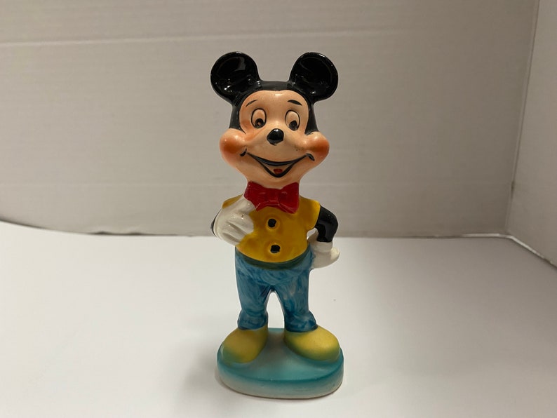 May include: A ceramic figurine of Mickey Mouse wearing a yellow shirt, blue pants, and a red bow tie. He is standing on a blue base.