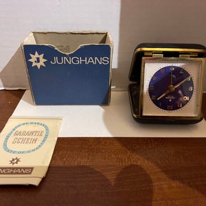 May include: A vintage Junghans travel alarm clock with a blue face and gold-tone accents, displayed open. The clock is in a black case, with a matching blue box and a guarantee certificate. The clock face has white numerals.