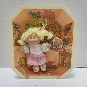 Puede incluir: Una muñeca vintage con coletas rubias, vestida con una blusa blanca y una falda rosa, dentro de una caja de plástico decorativa. La caja tiene un interior pintado con ilustraciones de juguetes y la palabra "MILK". La muñeca sostiene un osito de peluche.