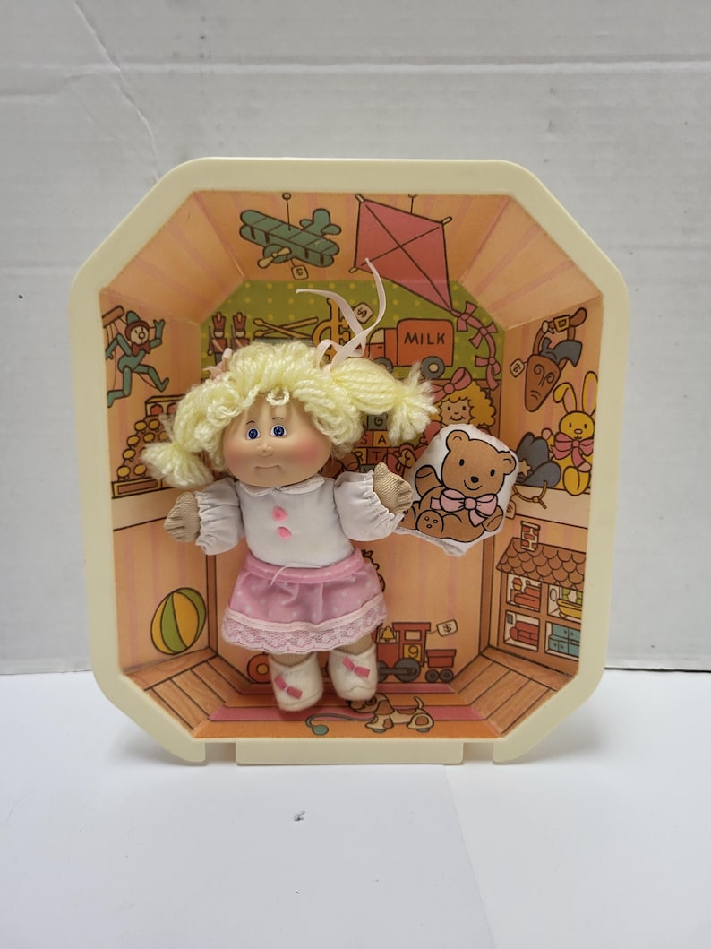 May include: A vintage doll with blonde pigtails, wearing a white top and pink skirt, inside a decorative plastic case. The case has a painted interior with toy-themed illustrations and the word "MILK". The doll holds a teddy bear.