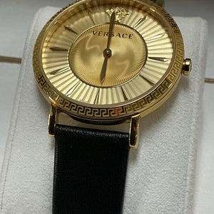 Pu&ograve; includere: Un orologio analogico color oro con cassa rotonda e motivo a chiave greca sulla lunetta. Il quadrante dorato mostra il logo Versace, un emblema con testa di Medusa a ore dodici e sottili lancette dorate, abbinato a un cinturino in pelle nera.
