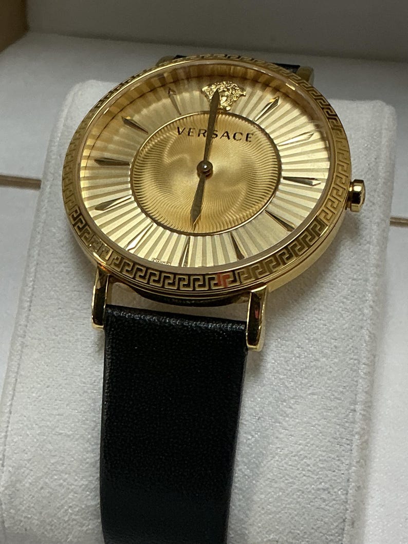 Pu&ograve; includere: Un orologio analogico color oro con cassa rotonda e motivo a chiave greca sulla lunetta. Il quadrante dorato mostra il logo Versace, un emblema con testa di Medusa a ore dodici e sottili lancette dorate, abbinato a un cinturino in pelle nera.