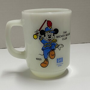 May include: White ceramic mug with a cartoon image of Mickey Mouse dressed as a bandleader. The mug has the text "The Mickey Mouse Club" and "1955" printed on it.