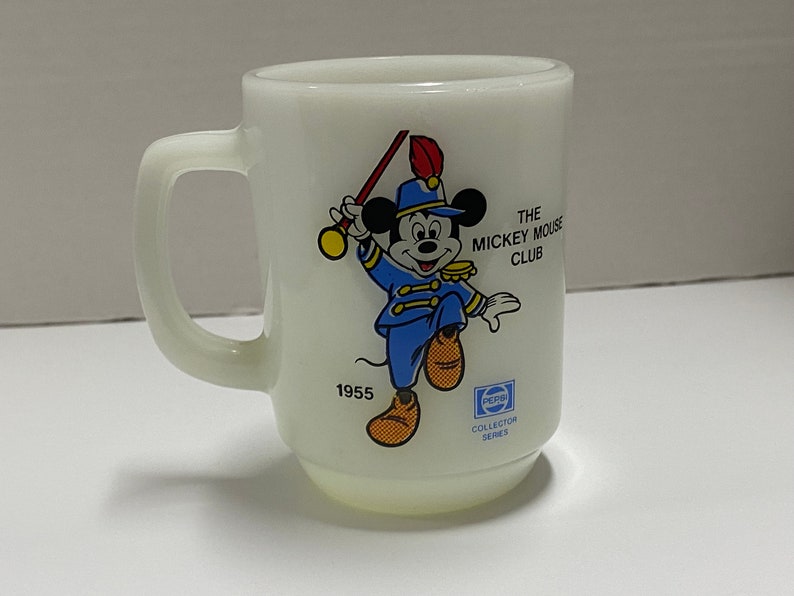 May include: White ceramic mug with a cartoon image of Mickey Mouse dressed as a bandleader. The mug has the text "The Mickey Mouse Club" and "1955" printed on it.