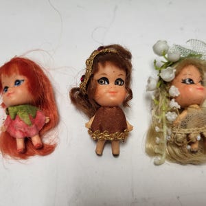 Könnte beinhalten: Drei Miniaturpuppen im Vintage-Stil. Die erste Puppe hat rotes Haar und ein rosa Kleid. Die zweite Puppe hat braunes Haar und ein braunes Kleid mit goldenen Verzierungen. Die dritte Puppe hat blondes Haar und ein beiges Kleid mit weißen Blumenakzenten.