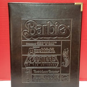 May include: A dark brown leather-bound binder with embossed text and logos. The binder features the Barbie logo, along with "Preschool SEE 'N SAY", "Mönchhichi", "MASTERS OF THE UNIVERSE", "Teach & Learn Computer", and "DRIVE COMMAND". Gold corner protectors are visible.