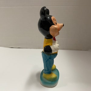 May include: A ceramic figurine of Mickey Mouse wearing a yellow vest, blue jeans, and a red bow tie. He is standing on a blue base.