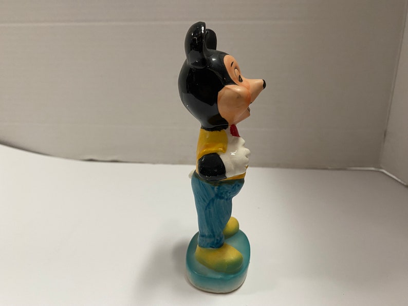 May include: A ceramic figurine of Mickey Mouse wearing a yellow vest, blue jeans, and a red bow tie. He is standing on a blue base.