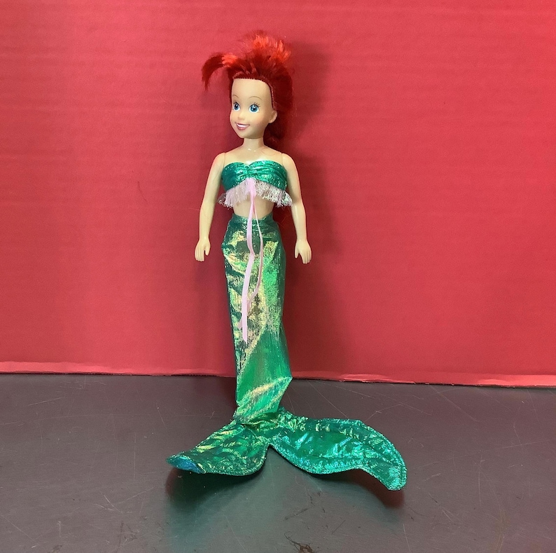 May include: A red-haired mermaid doll with a green tail and a pink and white top. The doll has a smiling face and is wearing a green and gold tail.