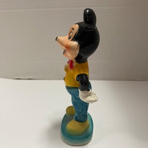 May include: A ceramic figurine of Mickey Mouse wearing a yellow shirt, blue pants, and a red bow tie. He is standing on a blue base.