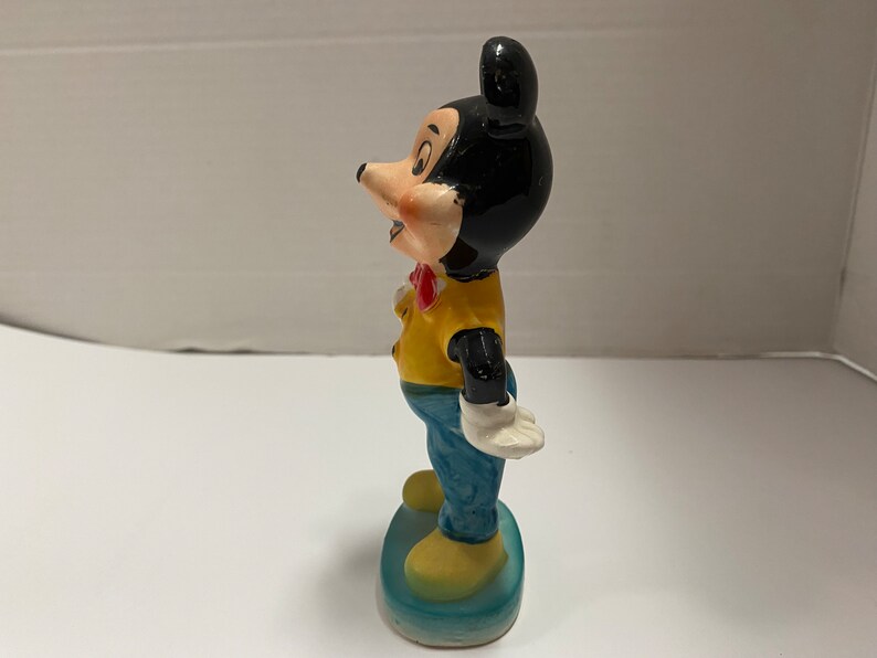 May include: A ceramic figurine of Mickey Mouse wearing a yellow shirt, blue pants, and a red bow tie. He is standing on a blue base.